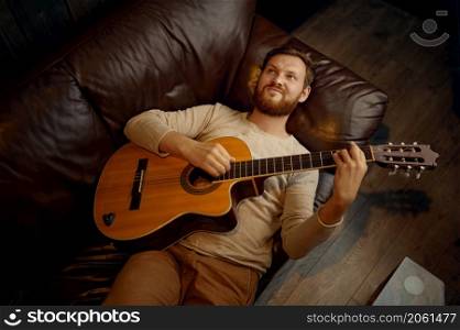 Dreamy smiling pensive romantic man lying on couch playing guitar looking up overhead view. Dreamy man lying on couch playing guitar
