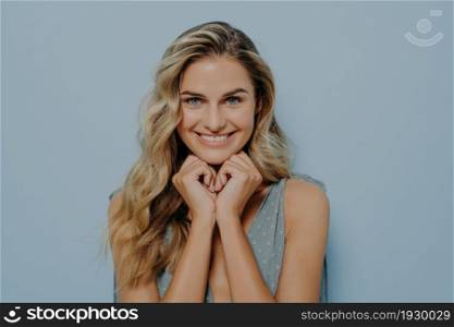 Dreamy young blonde woman in blue dress with hands pressed together under chin, rejoicing after hearing heartwarming complimentary words from stranger, isolated over blue background. Dreamy young blonde woman with hands pressed together under chin
