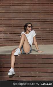 Dreamy young slender woman in trendy jean shorts and sunglasses chilling on wooden stage in summer and looking away. Slender female relaxing on tribune