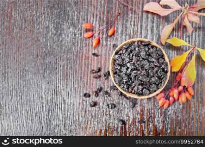Dried black barberry in a bowl, sprigs with fresh berries and leaves on dark wooden board background from above