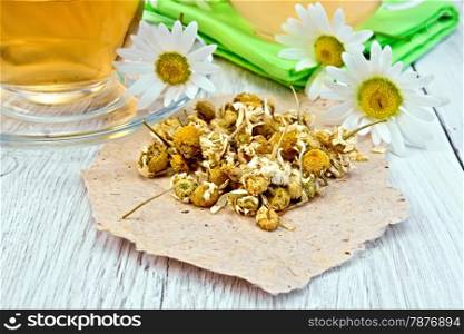 Dried chamomile flowers on paper, tea in glass teapot on napkin and cup, fresh chamomile flowers on background of wooden boards