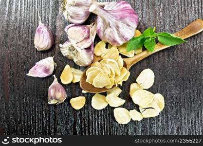 Dried garlic slices in a spoon and on the table, fresh garlic, basil on background of wooden board from above