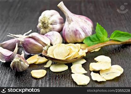 Dried garlic slices in a spoon and on the table, fresh garlic, basil on dark wooden board background