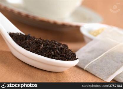 Dried loose black tea on ceramic spoon with tea bags, brown sugar and a cup in the back photographed on wood (Selective Focus, Focus one third into the leaves on the spoon)