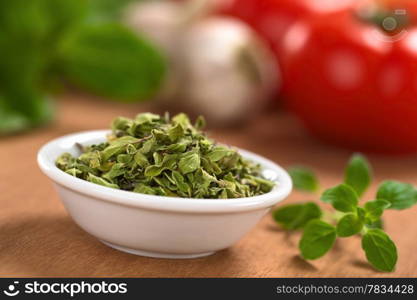 Dried oregano leaves in small bowl with fresh oregano on the side, tomato, garlic and basil in the back (Selective Focus, Focus one third into the dried oregano leaves). Dried Oregano