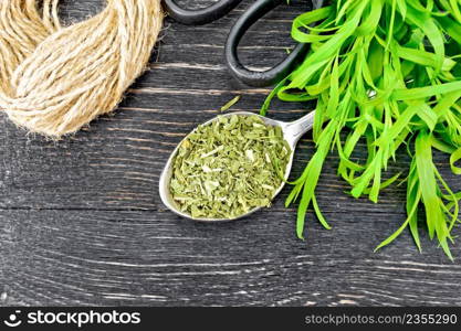 Dried tarragon in a spoon, fresh estragon, a skein of twine and scissors on black wooden board background from above