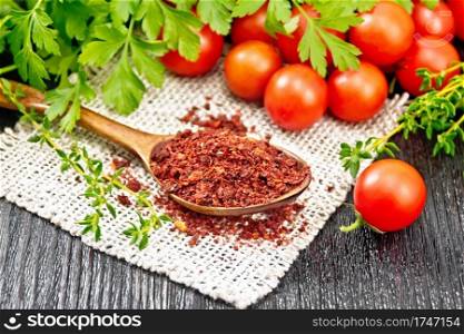 Dried tomato flakes in a spoon on burlap, fresh small tomatoes, parsley and thyme on black wooden board background