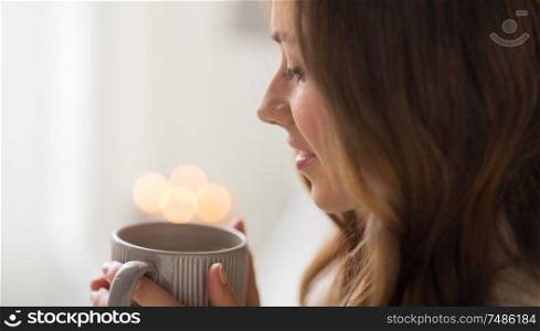 drink, christmas and hygge concept - close up of happy woman with lights over cup of coffee or tea at home. close up of happy woman with cup of coffee at home