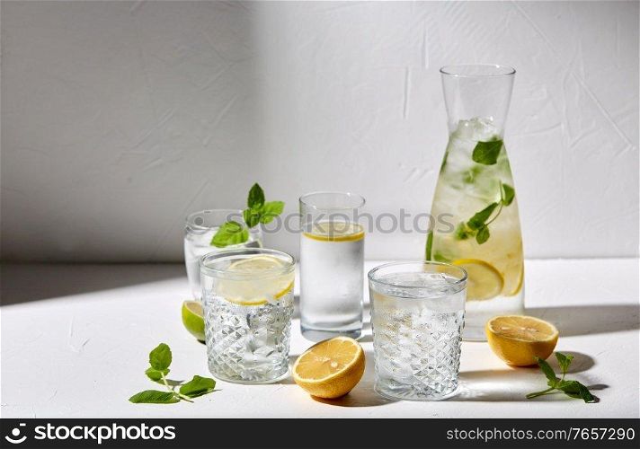 drink, detox and diet concept - glasses with fruit water or lemonade, lemons, limes and peppermint on white table. glasses with lemon water and peppermint on table