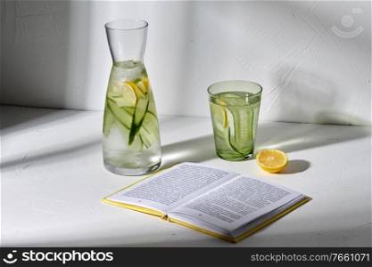 drink, detox and diet concept - glasses with fruit water with lemon and cucumber and open book dropping shadows on white surface. glass of water with lemon and cucumber and book