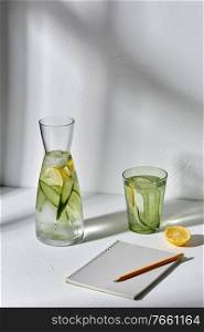 drink, detox and diet concept - glasses with fruit water with lemon and cucumber and notebook with pencil dropping shadows on white surface. water with lemon and cucumber and notebook