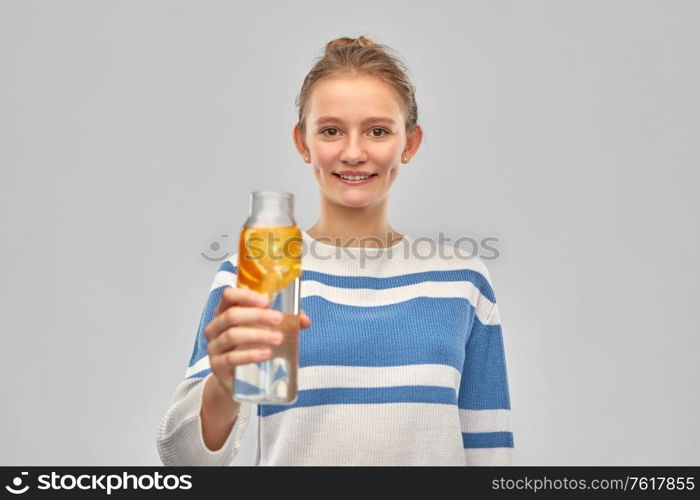drink, health and people concept - happy smiling teenage girl in sweater with orange fruit infused water in glass bottle over grey background. teenage girl with glass bottle of fruit water