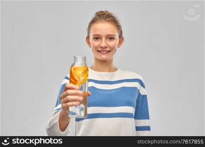 drink, health and people concept - happy smiling teenage girl in sweater with orange fruit infused water in glass bottle over grey background. teenage girl with glass bottle of fruit water