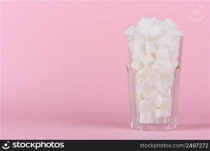 Drinking glass of of lump sugar cubes on pink pastel background. Unhealthly diet with sweet sugary soft drinks concept.
