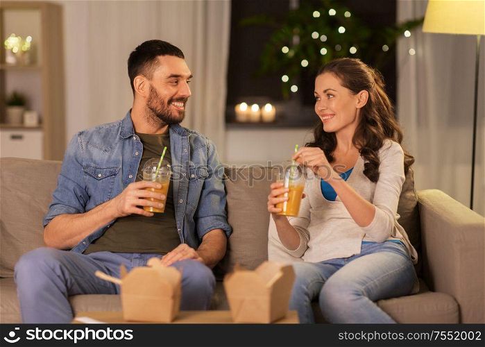 drinks and people concept - happy couple drinking takeaway juice from plastic cups with paper straws at home in evening. happy couple drinking takeaway juice at home