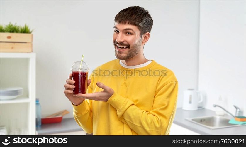 drinks and people concept - happy smiling young man holding tomato juice in takeaway plastic cup with paper straw over home kitchen background. happy man with tomato juice in takeaway cup