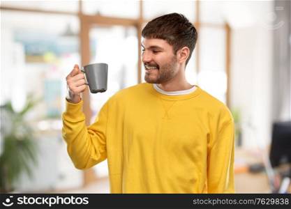 drinks and people concept - happy smiling young man with coffee cup over office background. happy smiling young man with coffee cup at office