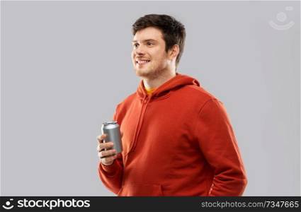 drinks and people concept - happy young man in red hoodie drinking soda from tin can over grey background. happy young man drinking soda from tin can