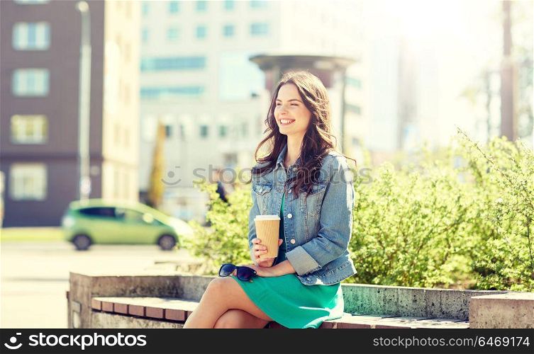 drinks and people concept - happy young woman or teenage girl drinking coffee from paper cup sitting on on city street bench. happy young woman drinking coffee on city street