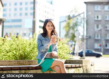 drinks and people concept - happy young woman or teenage girl with handbag drinking coffee from paper cup sitting on on city street bench. happy young woman drinking coffee on city street