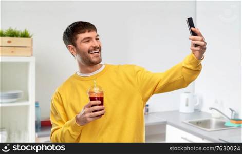drinks and people concept - smiling man taking selfie with smartphone with tomato juice in takeaway plastic cup over home kitchen background. happy man with smartphone and juice taking selfie