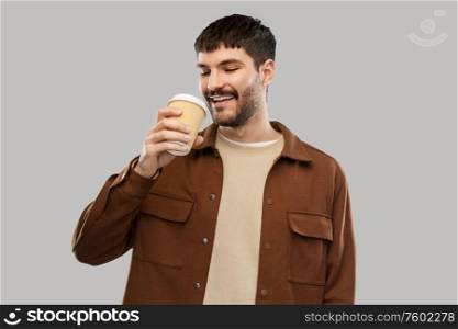 drinks and people concept - smiling young man drinking coffee from takeaway cup over grey background. happy young man drinking coffee from takeaway cup