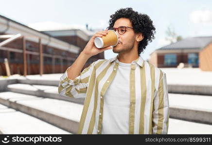 drinks and people concept - young man in glasses drinking takeaway coffee from paper cup over city street background. man drinking coffee from paper cup in city