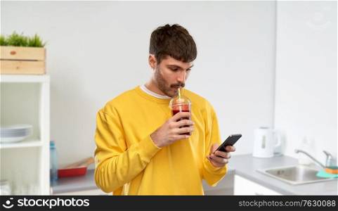 drinks and people concept - young man with smartphone and tomato juice in takeaway plastic cup with paper straw over home kitchen background. man with smartphone and tomato juice