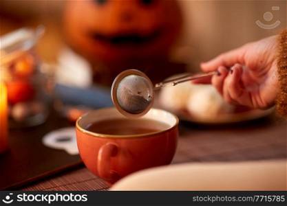 drinks, holidays and leisure concept - close up of woman’s hand with mesh tea infuser ball and mug at home on halloween. woman’s hand with tea infuser and mug on halloween