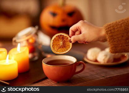drinks, holidays and leisure concept - woman’s hand adding dry orange slice to cup of tea at home on halloween. hand adding dry orange to cup of tea on halloween