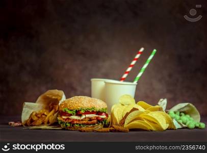 Drinks in a paper cup with a paper tube and hamburger, salty snacks, pretzels, chips.. Burger on a rustic wooden board and unhealthy snacks and chips, drinks in a paper cup with a paper tube