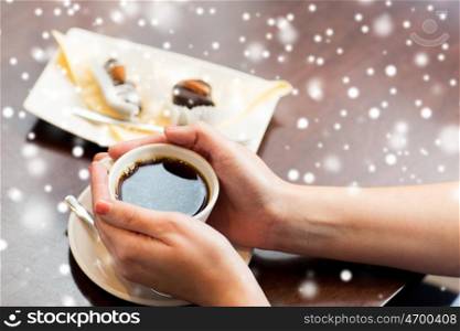 drinks, people and winter holidays concept - close up of woman hands holding cup with black coffee and dessert over snow