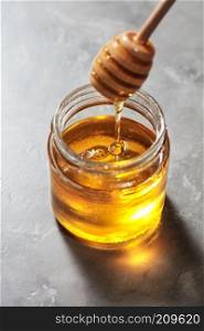 Dripping sweet natural organic honey from stick into glass jar on a gray marble background, pure natural raw sweet goodness.. Natural sweet golden dripping honey in a glass pot with dipper on a gray stone table. Rosh hashanah jewish holiday.