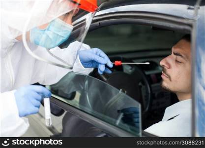 Drive-thru Coronavirus COVID-19 test,paramedic in personal protective equipment & face shield performing nasal swab through car window on male patient sitting in vehicle,mobile US testing site center