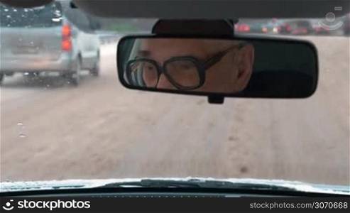 Driving in the city in winter. Senior man in glasses driving on messy road, his face reflecting in rear mirror