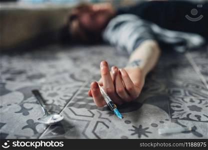 Drug addict lies on the floor, hand with syringe and spoon for dose preparing on the table. Addiction concept, addicted people. Drug addict, syringe and spoon for dose preparing