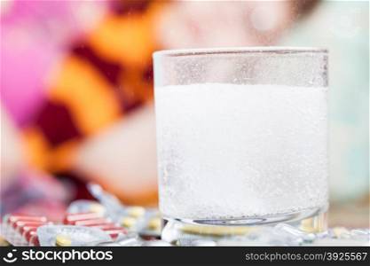 drug dissolves in glass with water and pills on table close up and sick woman with scarf around her neck on sofa in living room on background