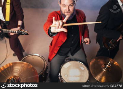 Drummer in red suit, performing on the stage of night club, electro and bas-guitarists on background. Rock band concert, music show