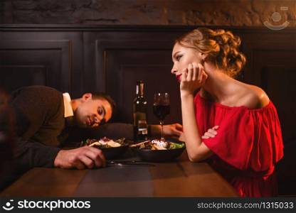 Drunk man sleeps at the table against woman in red dress in restaurant. Couple have a spoiled evening. Drunk man sleeps at the table against woman