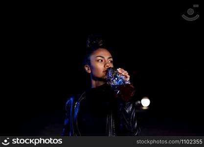 drunk young African American mulatto girl in a leather jacket and black clothes is on an abandoned sandy road drinking from a bottle of whiskey. at night in the light of car headlights and street lights