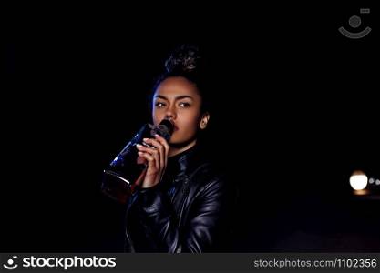drunk young African American mulatto girl in a leather jacket and black clothes is on an abandoned sandy road drinking from a bottle of whiskey. at night in the light of car headlights and street lights