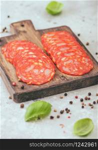 Dry-cured chorizo sausage slices with a distinctive spicy paprika flavour with basil and pepper on wooden chopping board and light background.