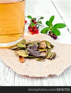 Dry lingonberry leaf on paper, tea in glass mug, berries and green leaves on a background of red bilberry bright wooden planks