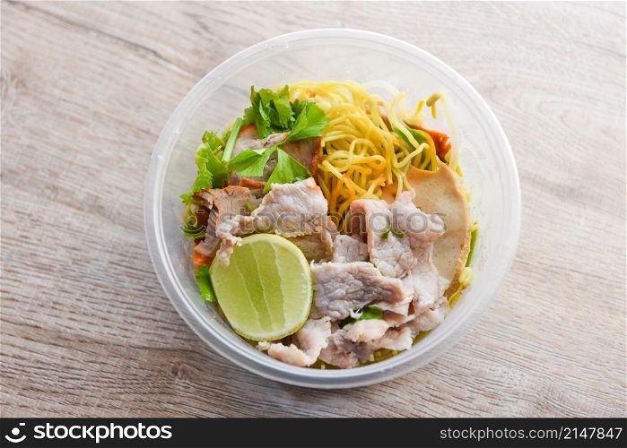Dry noodles pork with red roasted pork and vegetable in plastic bowl on wooden table, top view Thai and China Asian food yellow noodles lemon lime