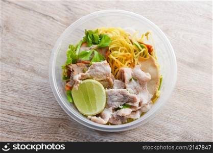 Dry noodles pork with red roasted pork and vegetable in plastic bowl on wooden table, top view Thai and China Asian food yellow noodles lemon lime