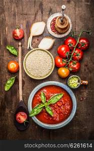 Dry Quinoa in ceramic bowl with tomatoes sauce and fresh ingredients for cooking, top view. Quinoa preparation on rustic wooden background.