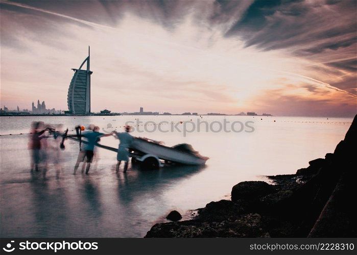 DUBAI, UAE - FEBRUARY 2018  The world’s first seven stars luxury hotel Burj Al Arab at sunset seen from Jumeirah public beach in Dubai, United Arab Emirates. People prepare a boat to go out fishing in the sea.