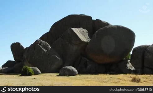 Dunkle runde Steingebilde, runde und kantige schwarze Steine liegen ubereinander. Sandiger Boden, blauer Himmel.
