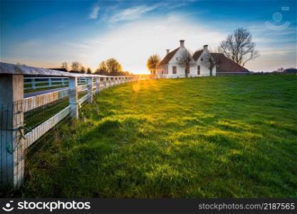Dutch landscape with historical houses in evening along a curved road with white fence in the country - Groningen, Holland, Europe. Country road in dutch countryside with white fence leading towards an old farmhouse in Holland during an atmospheric sunset