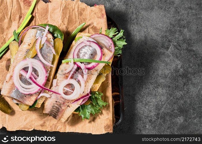 Dutch sandwich with herring, pickled cucumbers and red onions on a paper lining. Ingredients on a cutting board nearby. Layout on the table, selective focus and copy space
